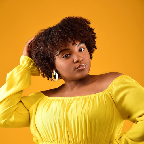 A model wearing a yellow shirt while touching her hair in front of an orange backdrop.